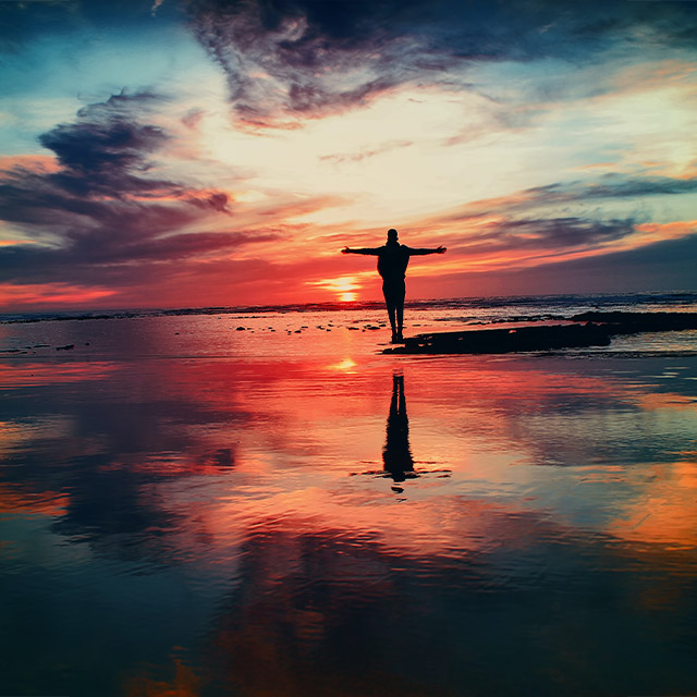 A silhouette stands on a beach at sunset, with vibrant colors reflecting on the water's surface and clouds scattered in the sky.