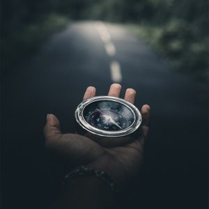 A hand holding a compass, with a dark, blurred road stretching ahead in the background.