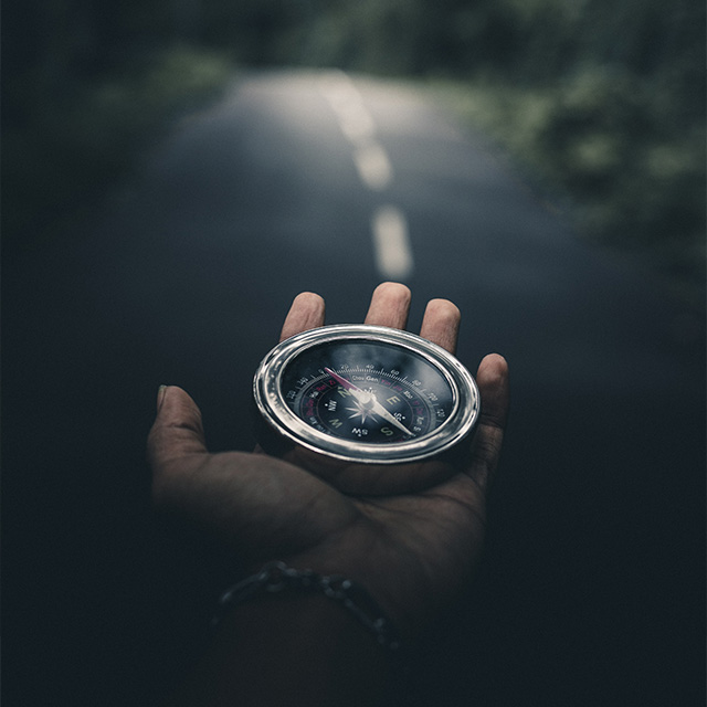 A hand holding a compass, with a dark, blurred road stretching ahead in the background.