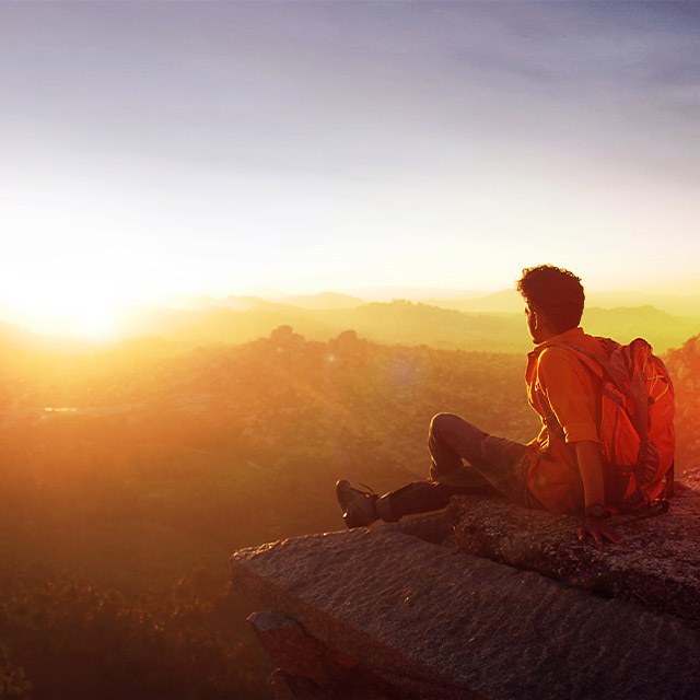 A person sits on a rocky ledge, gazing at a warm sunset over distant mountains.