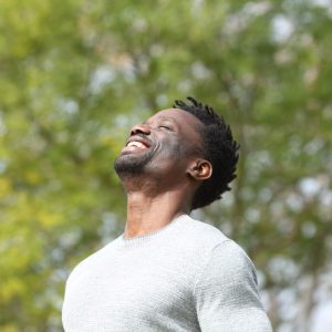 A man with a joyful expression stands outdoors, basking in the sunlight, with greenery in the background.